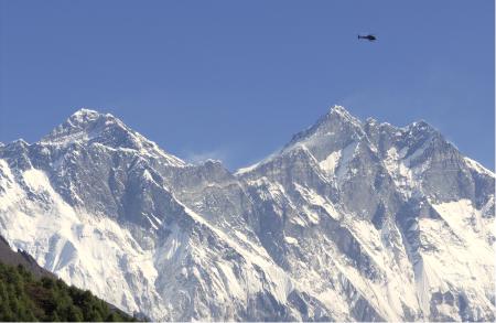 The snowclad peaks of Mount Everest and Lhotse, with patches of black rock visible. A helicopter flies by them.