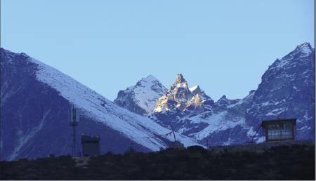 A telecom tower stands in front of a snowy rocky pinnacle in the mountainous landscape.