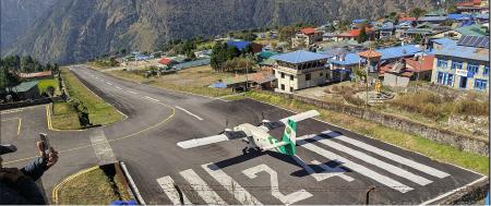 The runway of Lukla Airport, seen from one end, with the runway sloped down leading to the ridge. A green and white Tara Air twin-otter plane waits on the high end
