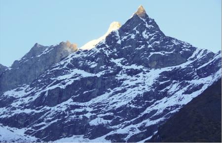 Sun rays strike the pointy peak of a mountain. Partially covered in snow, the mountain's dark rock surface is visible.