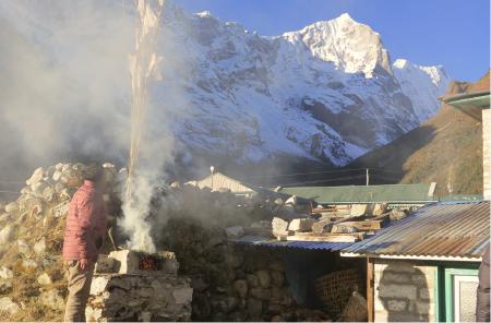A person stands beside rising incense smoke from a pedestal, as sunlight hits the peak of a snowcapped mountain.