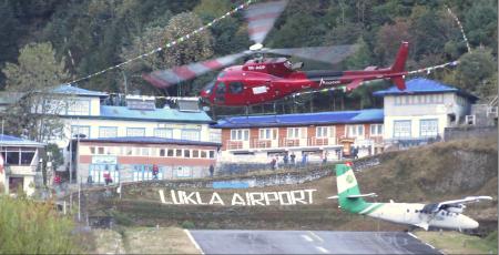 A red helicopter tilts sideways while approaching the helipad to land, in the forground of a small hill with giant white letters spelling Lukla Airport. A green and white Tara Air twin-otter plane is taxing to the side on the same runway.