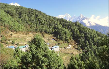 A small village on a mountain, with a few houses scattered along the hillside. Three mountain peaks stand in the background