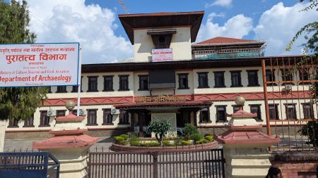 The Department of Archaeology and National Archives Building: A long, two story building, with beige walls, carved wooden exterior window trims, and a protruding entrance area supported by carved wooden pillars