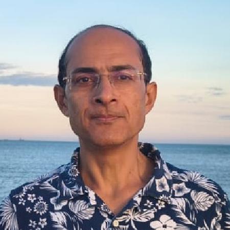 A portrait image of Neel Kamal Chapagain, in the foreground of an ocean at dusk. He is wearing a deep-blue shirt