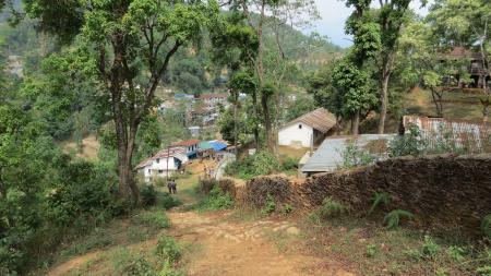 An image from the top of a green hill, facing a dusty sloped path downhill. At the foot of the slope are a few people, and beside the path are a few small houses. In the backdrop is the face of another mountain