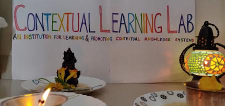 A banner with the title "Contextual Learning Lab" followed by "An Instituition For Learning & Promoting Contextual Knowledge Systems". In the foreground is a lamp, a type of candle that's lit, and a small statue